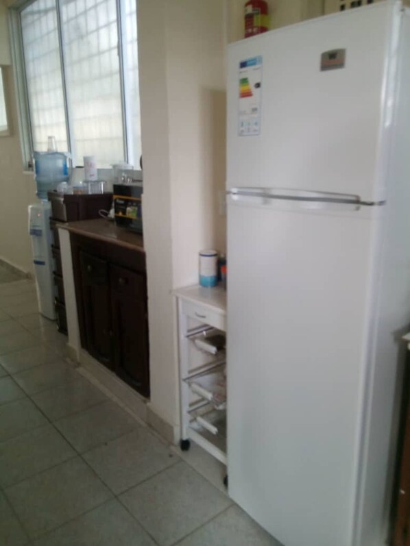 A kitchen with a white refrigerator, a water dispenser, and a microwave. There is a window with blinds and a cabinet with a shelf. - Hostal Arboleda, Colonial Zone, Santo Domingo (Santo Domingo)