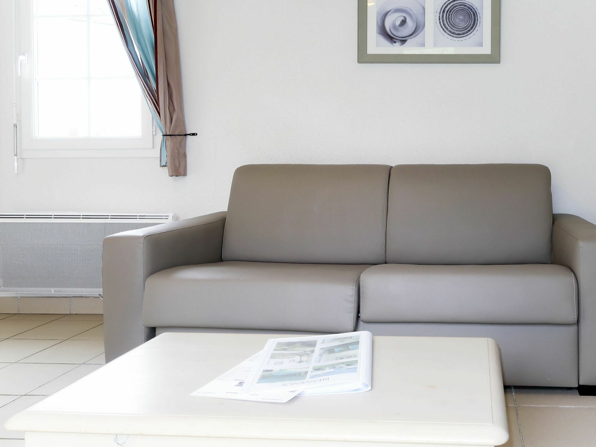 A modern living room with a beige sofa, a white coffee table with magazines, a radiator, and a window with curtains.