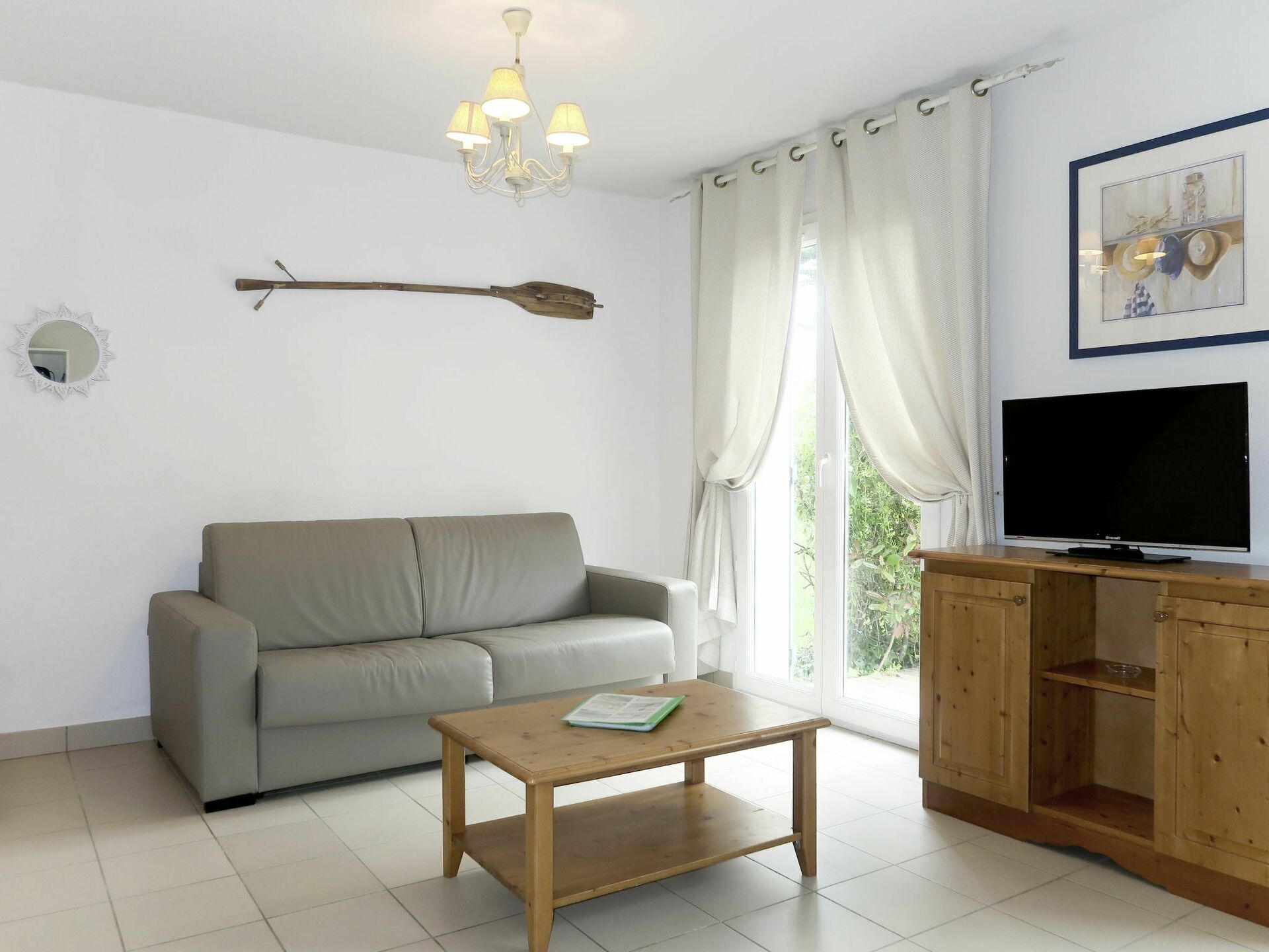 A living room with a sofa, a wooden coffee table, a TV on a wooden cabinet, a chandelier, and a window with curtains.