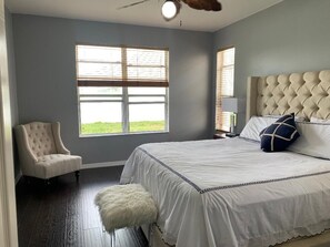 Ein Schlafzimmer mit einem großen Bett, einem gepolsterten Kopfteil, einem Sessel und einem Fenster mit Jalousien.