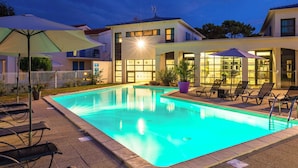 Una piscina con sillas de descanso y sombrillas, rodeada de edificios con grandes ventanales y un exterior bien iluminado.