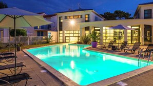 A swimming pool with lounge chairs and umbrellas, surrounded by buildings with large windows and a well-lit exterior. - Nice apartment for 6 people with pool, WIFI, hot tub, TV, balcony and pets allowed (Puyraveau)