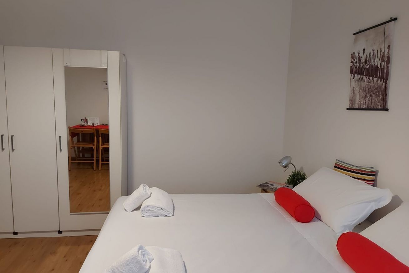 A hotel room with a bed, a wardrobe, a desk with a chair, and a small table with a lamp.