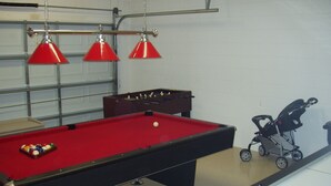 Taula de bitlles amb il·luminació vermella, taula de futbolí i un cotxet infantil en una habitació de parets blanques i porta de garatge.