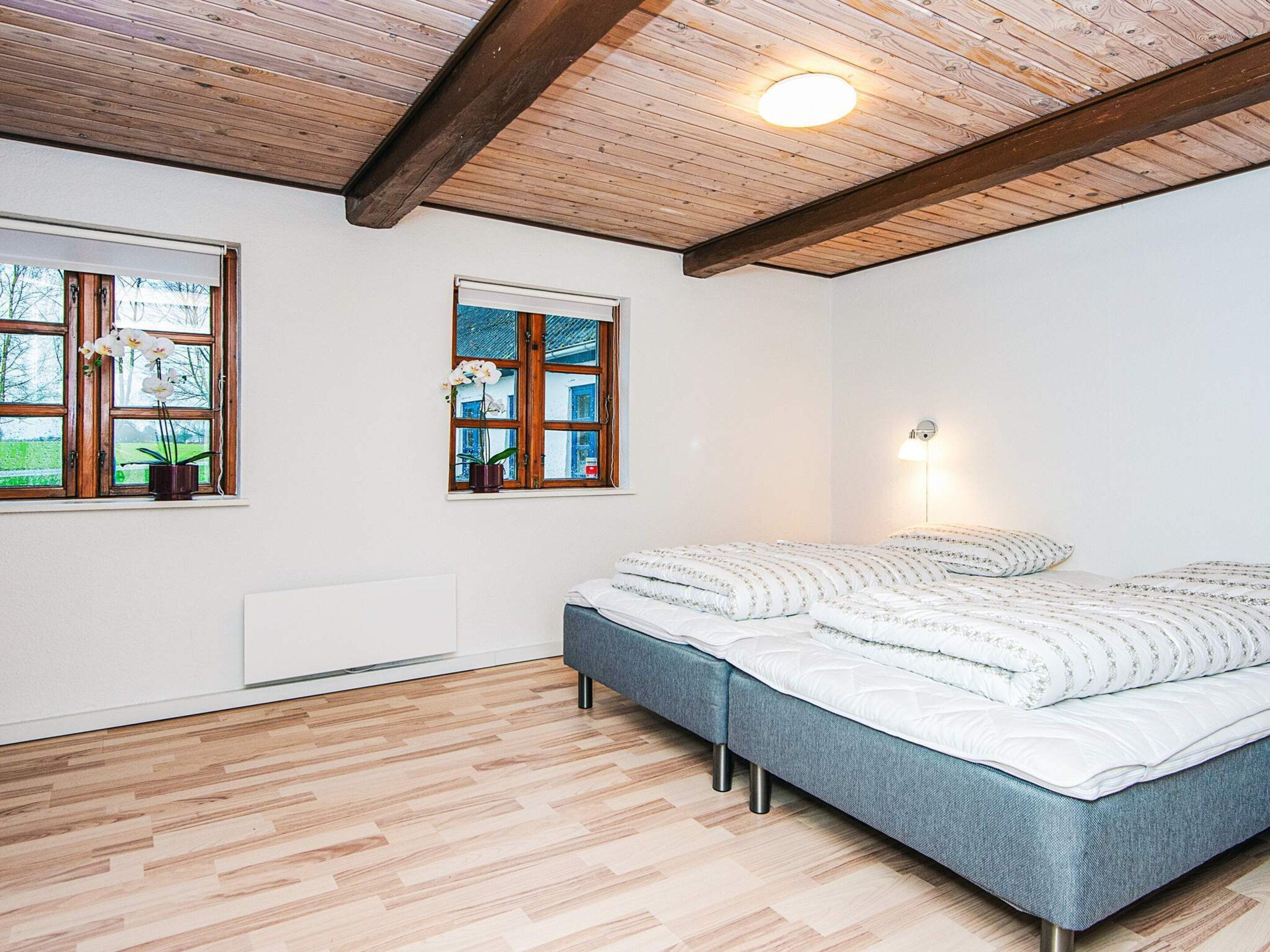 A bedroom with a bed, wooden flooring, a ceiling with exposed beams, and two windows with potted orchids.