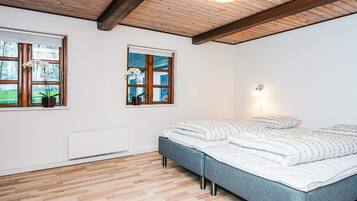 A bedroom with a bed, wooden flooring, a ceiling with exposed beams, and two windows with potted orchids.
