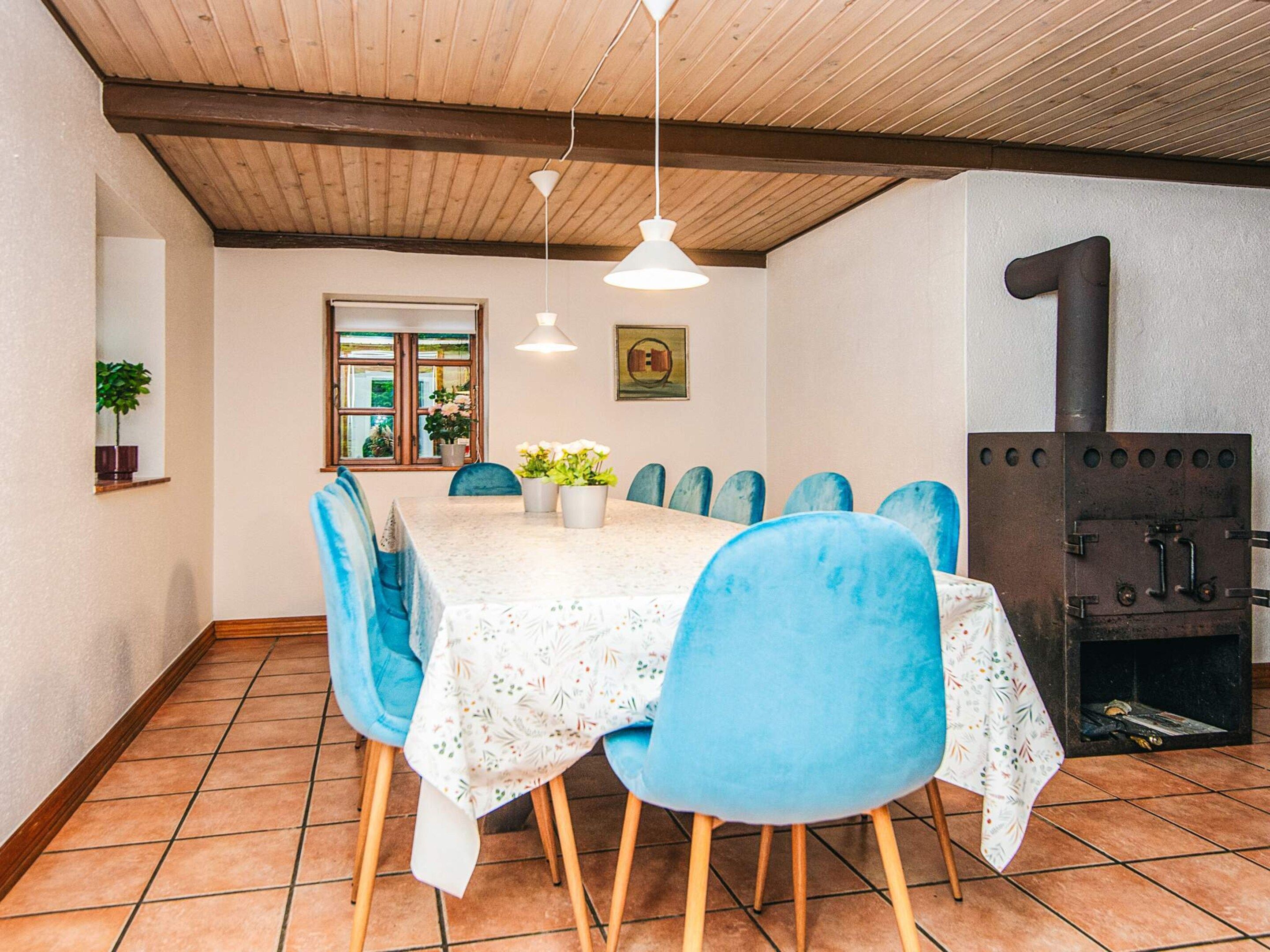 A dining area with a table covered in a floral tablecloth, surrounded by blue chairs, a wooden ceiling, and a rustic stove.