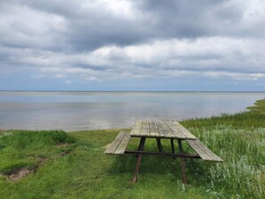 Una mesa de picnic de madera en un área verde al lado de un cuerpo de agua tranquilo, bajo un cielo nublado.