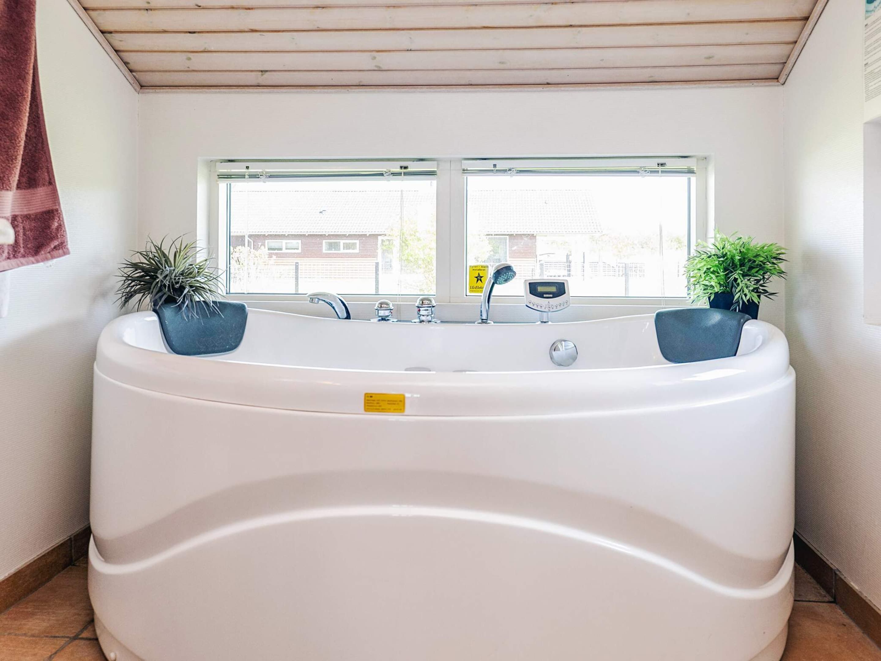 A bathroom with a large bathtub, two potted plants, a window with a view of buildings, and a towel hanging on the wall.