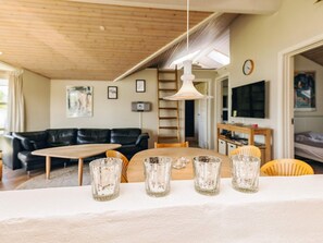A modern living room with a wooden ceiling, a black leather sofa, a wooden dining table with four clear glasses, and a television on the wall. - 5 Star Holiday Home in Rodby (Rodby)