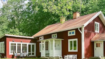 Una casa de madera roja con techo de tejas, un porche con barandillas blancas y un pequeño jardín con mobiliario exterior.