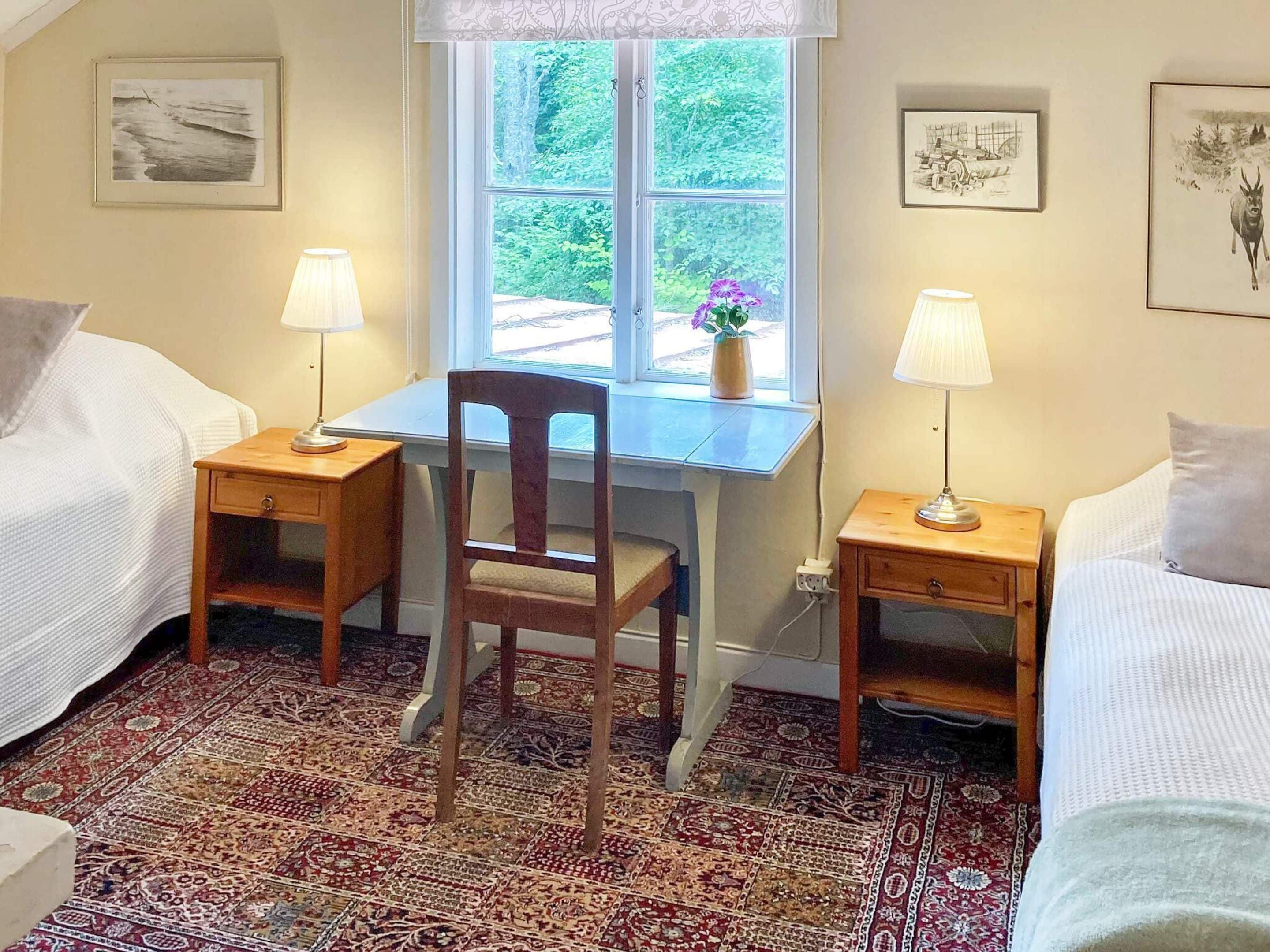 Een kamer met twee eenpersoonsbedden, een houten bureau met stoel, een raam met uitzicht op groen, en een kamerbreed tapijt met dessin.