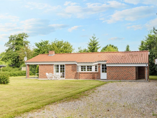A single-story brick house with a red-tiled roof, a covered patio with outdoor furniture, and a gravel driveway.