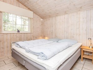 A bedroom with a wooden ceiling, a bed with white bedding, a nightstand with a lamp, and a window with a view of trees. - 4 Star Holiday Home in Orsted (Ørsted)