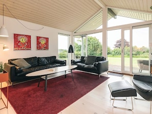 Een moderne woonkamer met een zwarte leren bank, een witte salontafel en een rood tapijt. Er zijn twee grote ramen met uitzicht op het groen.