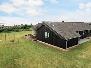 A black building with a sloped roof, a patio with a table and chairs, and a swing set in a grassy yard. - 5 Star Holiday Home in Faaborg (Faaborg)