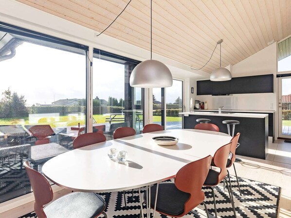 A modern kitchen with a large island, a dining area with a round table and chairs, and large windows offering a view of the outdoors.