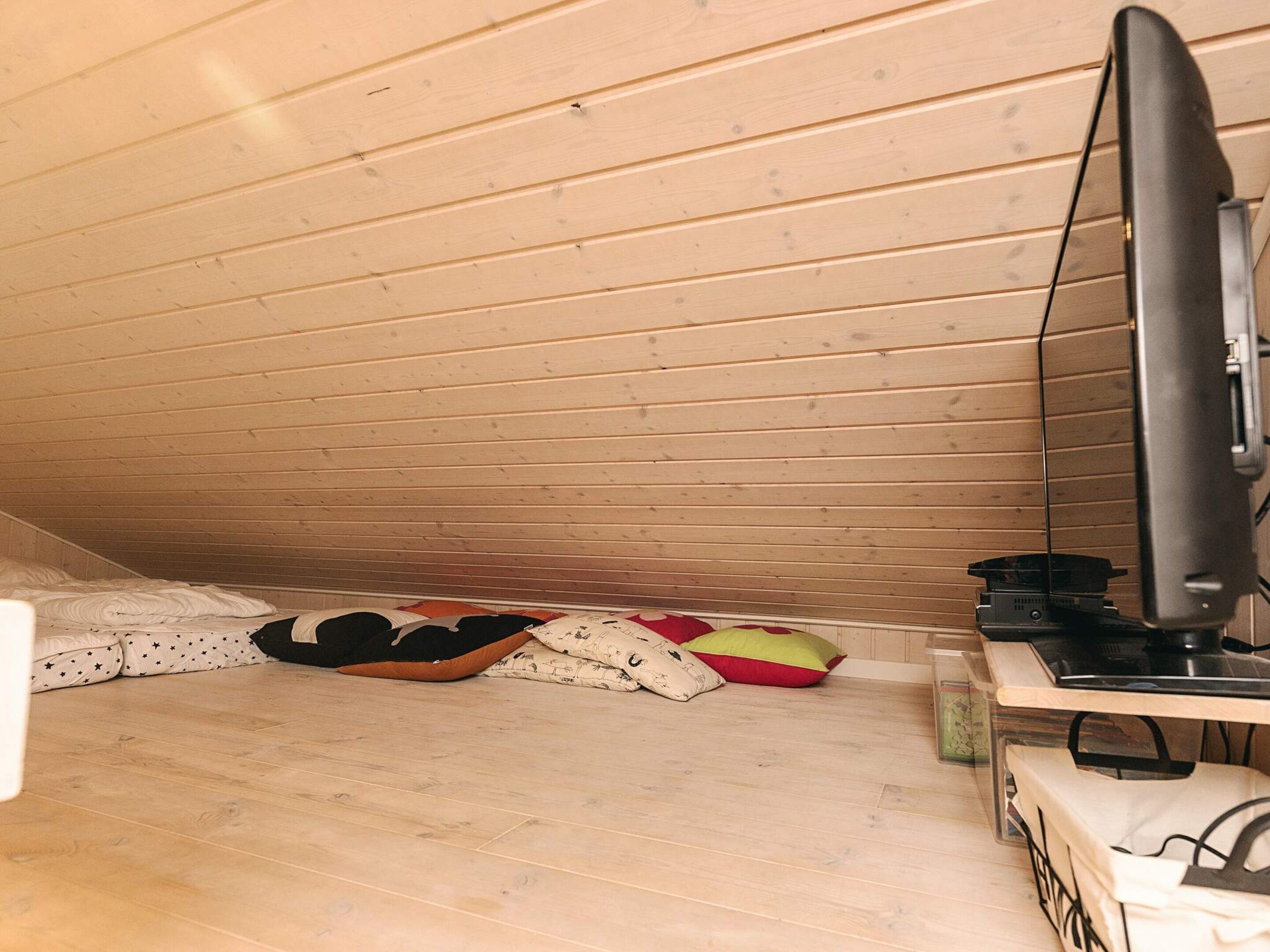 A small attic room with wooden slats on the ceiling, a television on a stand, and a few pillows on the floor.
