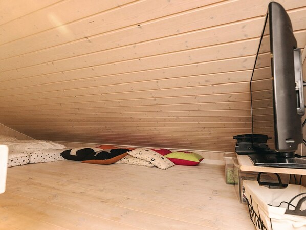 Une petite pièce mansardée avec des lambris en bois au plafond, un téléviseur posé sur un meuble et quelques coussins sur le sol.