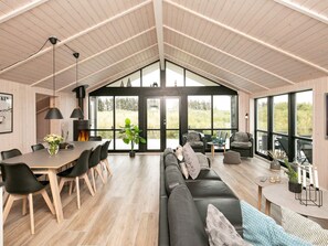 A spacious interior with a high ceiling, a dining area with a wooden table and chairs, a black leather sofa, and large windows offering a view of the outdoors.