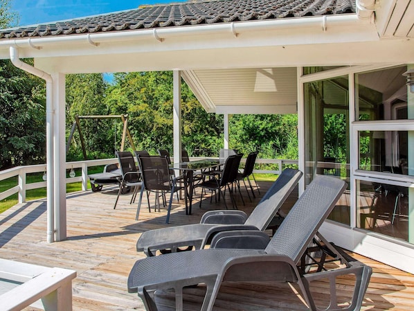 Katettu ulkoterassi, jossa on pöytä ja tuoleja, ja jota ympäröi valkoinen kaide. Terassilta on näkymä puutarhaan.