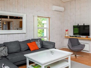 A living room with a grey sofa, white coffee table, and a grey armchair. There is a television on a wooden shelf, a window with curtains, and an air conditioning unit on the wall. - 4 Star Holiday Home in Vaeggerlose (Vaeggerlose)