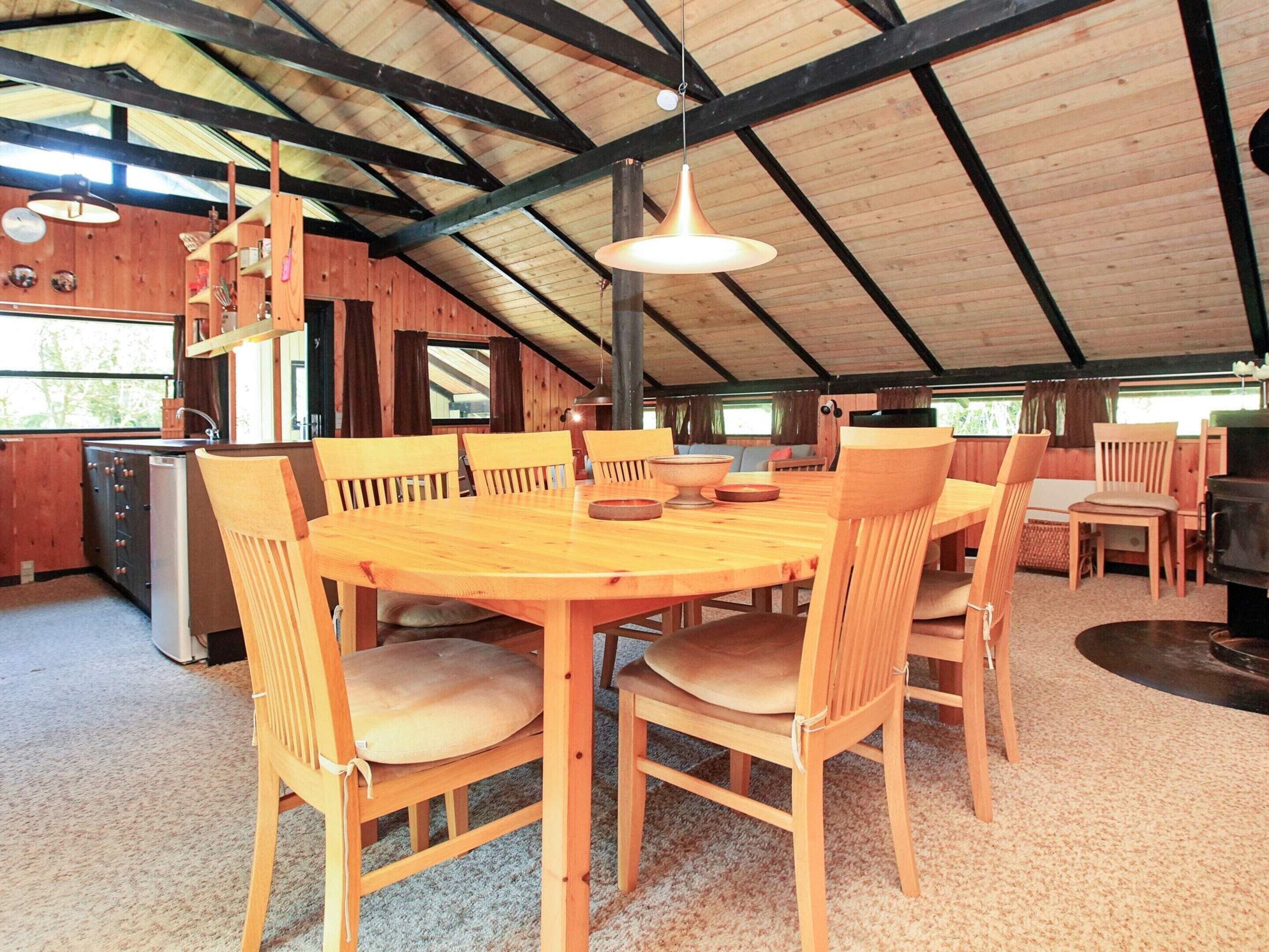 A wooden dining area with a large round table and chairs, a hanging light fixture, and a kitchen area with a grill.