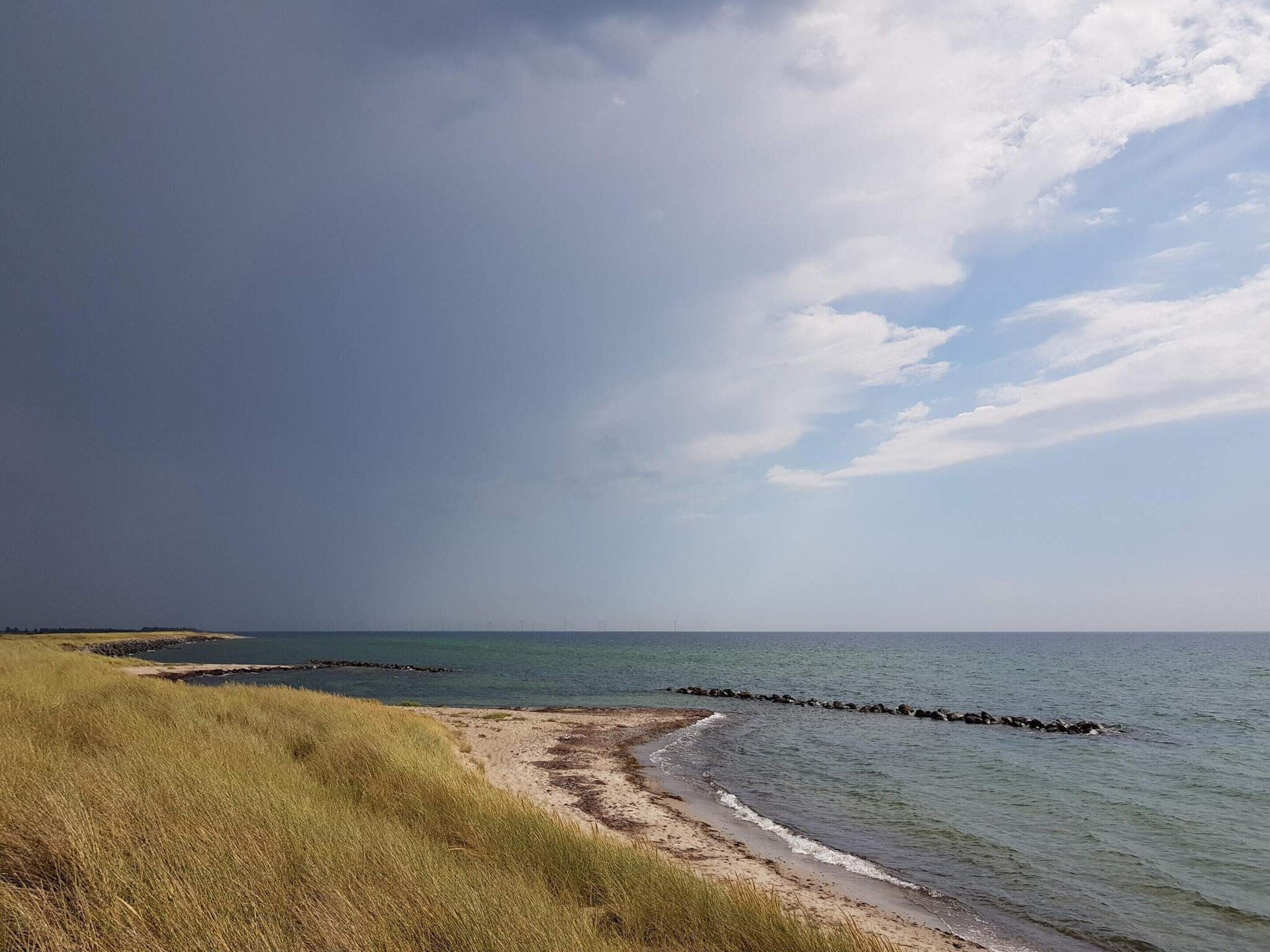 A beach with a grassy foreground, a rocky breakwater, and a calm sea under a partly cloudy sky.