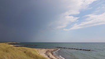 Ein Strand mit einer grasbewachsenen Vordergrund, einem steinernen Wellenbrecher und einem ruhigen Meer unter einem teils bewölkten Himmel.