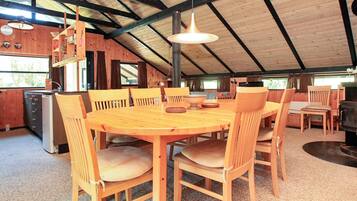 A wooden dining area with a large round table and chairs, a hanging light fixture, and a kitchen area with a grill.
