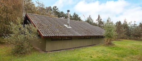 A small, single-story building with a sloped roof and a chimney, surrounded by a grassy field and trees.