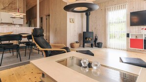 Ein modernes Interieur mit Holzbalkendecke, einem schwarzen Kaminofen, einem Essbereich mit Stühlen, einem an der Wand montierten Fernseher und einem Glastisch.