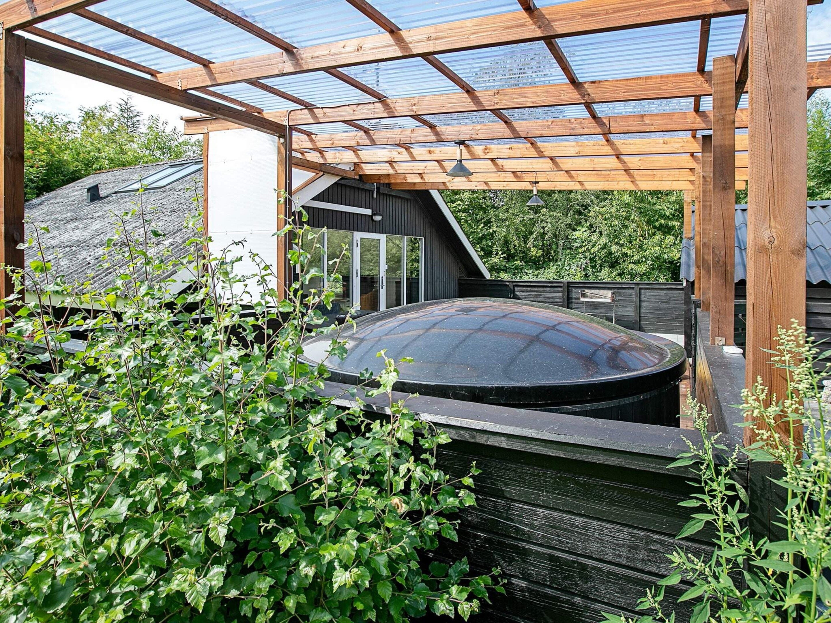 A hot tub under a wooden pergola with a glass ceiling, surrounded by greenery.