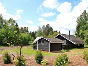 A two-story wooden cabin with a sloped roof, surrounded by a grassy yard and dense trees.