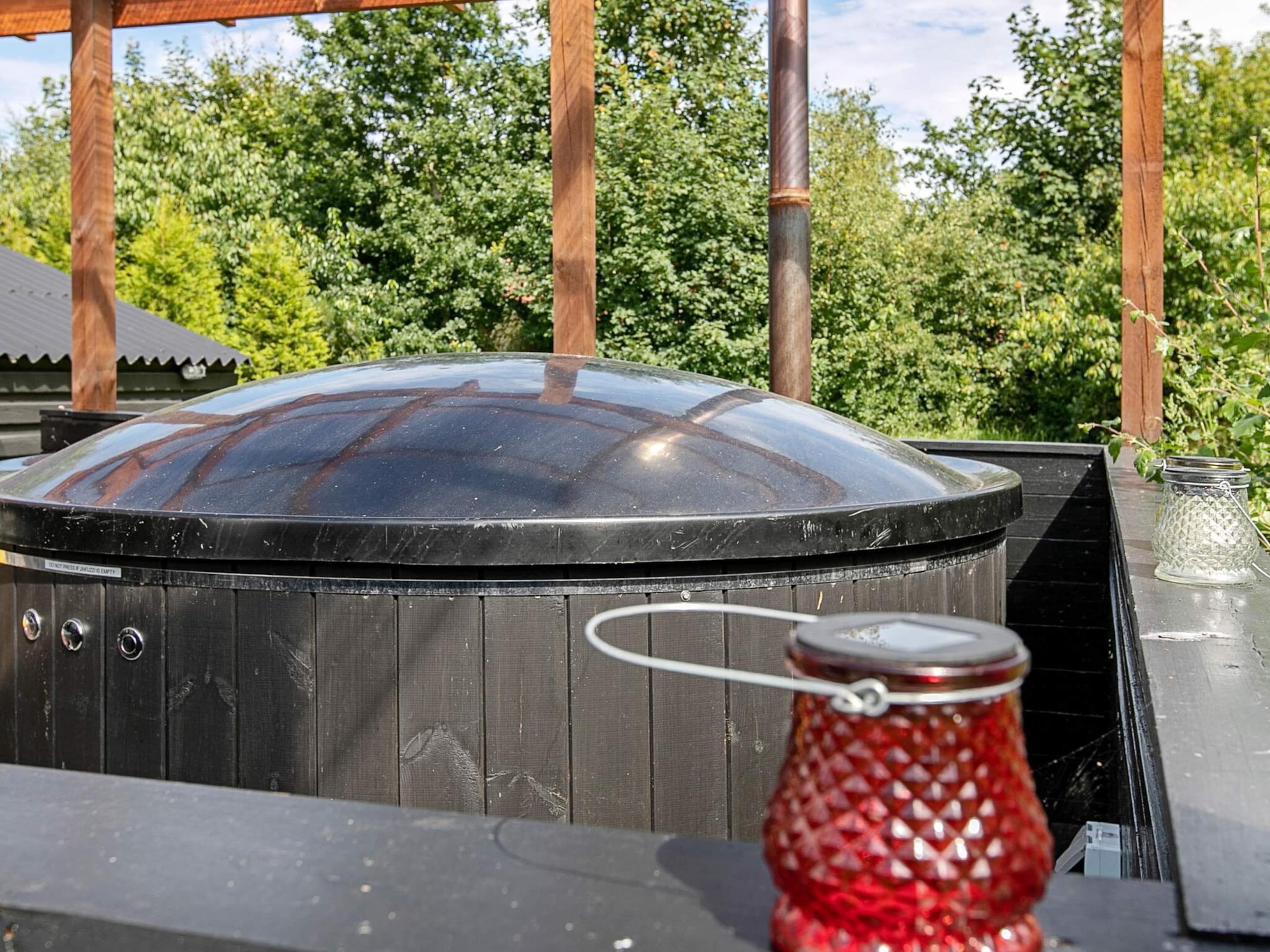 Et spabad med glaslåg, en rød dekorativ genstand og en metalkande på en træterrasse.