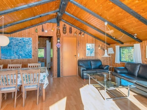 A wooden cabin interior with a dining area, a living room with a sofa and a glass coffee table, and a wooden ceiling with exposed beams.