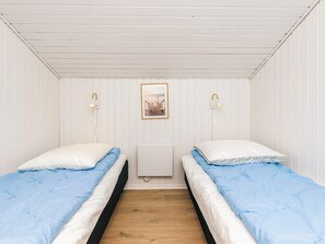 Une chambre avec deux lits, chacun recouvert d’une couverture bleue et agrémenté d’oreillers blancs, un plancher en bois, des murs blancs et un tableau encadré accroché au mur.