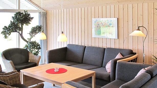 Um quarto acolhedor com um sofá cinzento, uma mesa de madeira, cadeiras de vime e uma parede de madeira com um quadro.