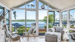 A spacious room with large windows offering a view of a lake and distant hills. There are two wooden chairs and a sofa with a fluffy throw. - 4 Star Holiday Home in Borkop (Borkop)