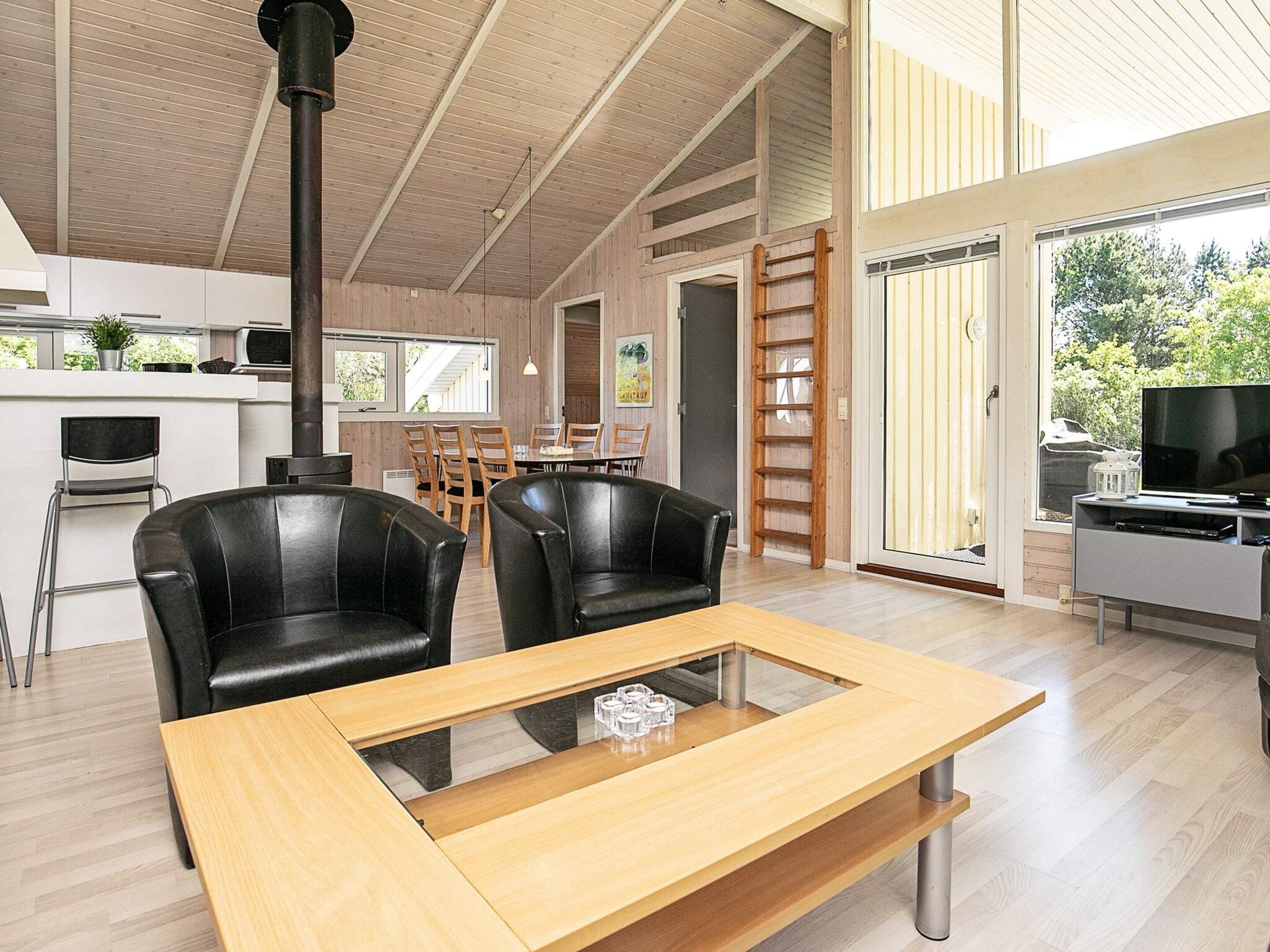 A modern living room with a glass-top coffee table, black leather chairs, a wooden dining table, and a large window offering a view of greenery.