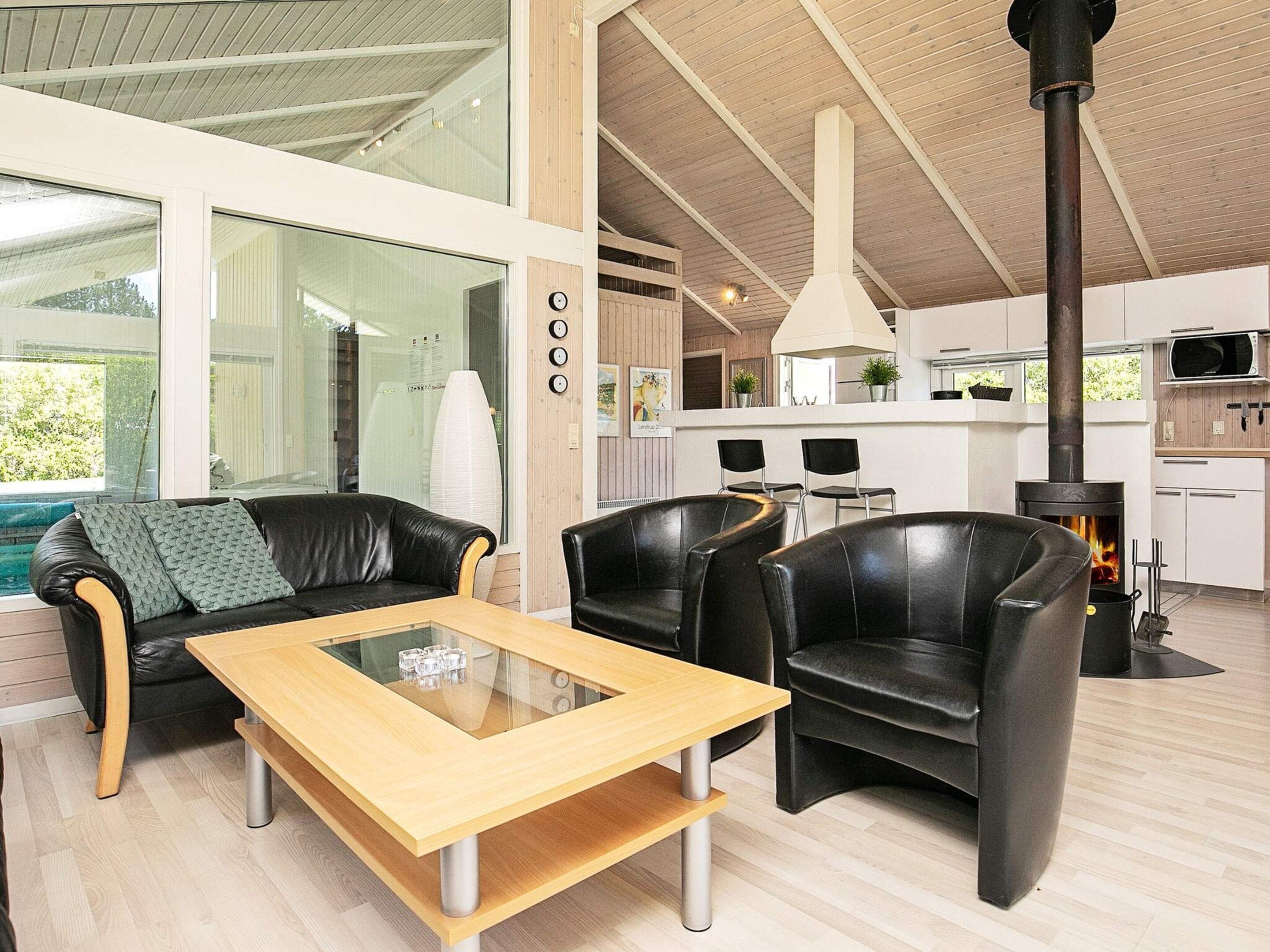 A modern interior with a glass-top coffee table, leather sofas, a kitchen area with a wood stove, and a view through large windows.