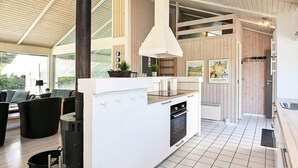 A modern kitchen with a central island, white cabinetry, and a tiled floor. There is a dining area with black furniture and a large window providing natural light. - 4 Star Holiday Home in Hjorring (Hjørring)