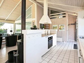 A modern kitchen with a central island, white cabinetry, and a tiled floor. There is a dining area with black furniture and a large window providing natural light.