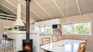A modern kitchen with a central wood-burning stove, a dining area with a round table and chairs, and a view of the outdoors through large windows. - 4 Star Holiday Home in Hjorring (Hjørring)