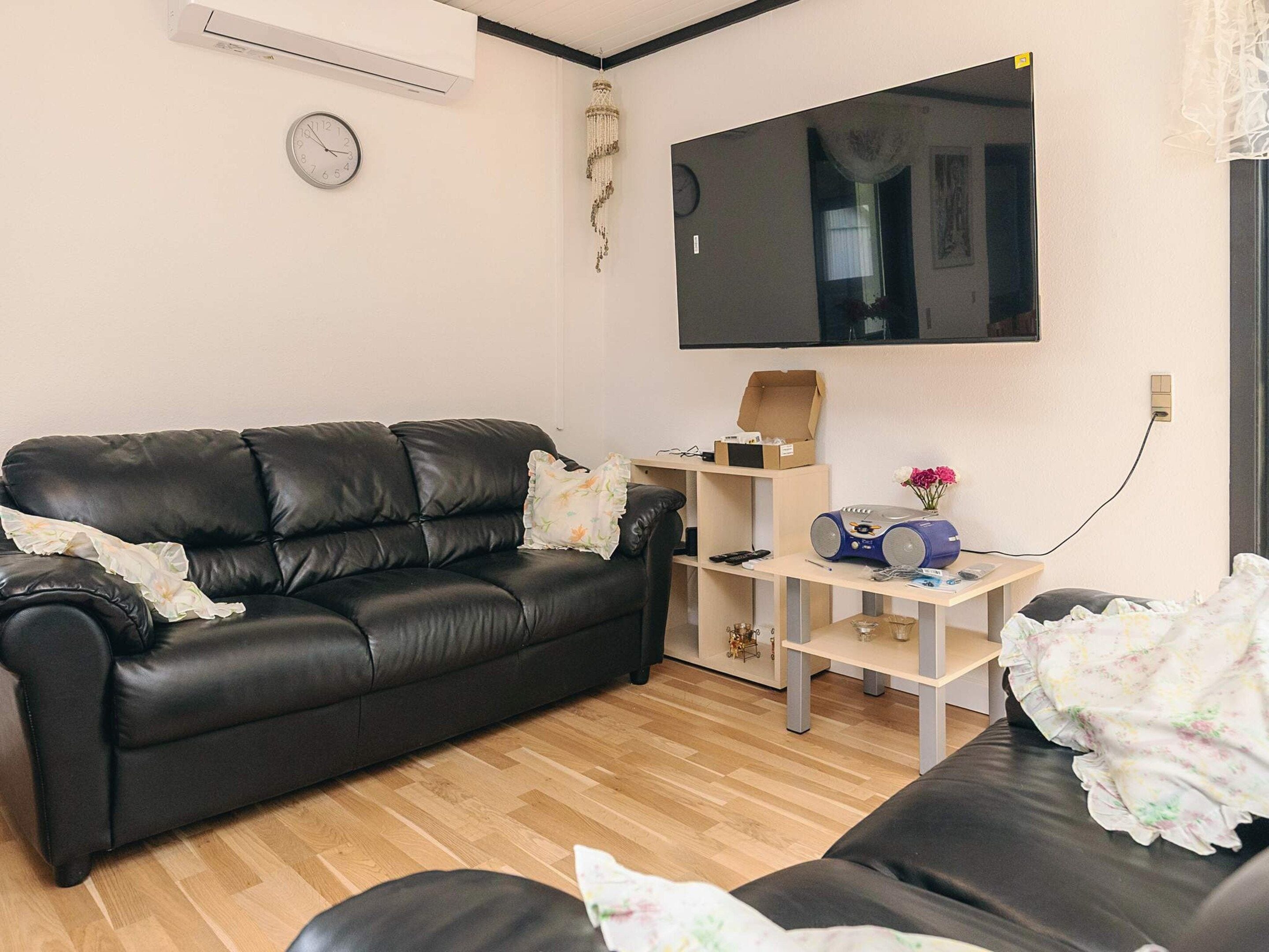 A living room with a black leather sofa, a wooden coffee table, a flat-screen TV mounted on the wall, and a clock on the wall.