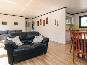 A living room with a black leather sofa, wooden dining table, and chairs. There are wall-mounted decorations and a kitchen area visible in the background.