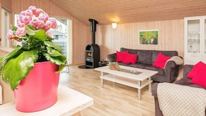 A cozy living room with a wooden ceiling, a sofa with red cushions, a coffee table, and a potted plant with pink flowers.