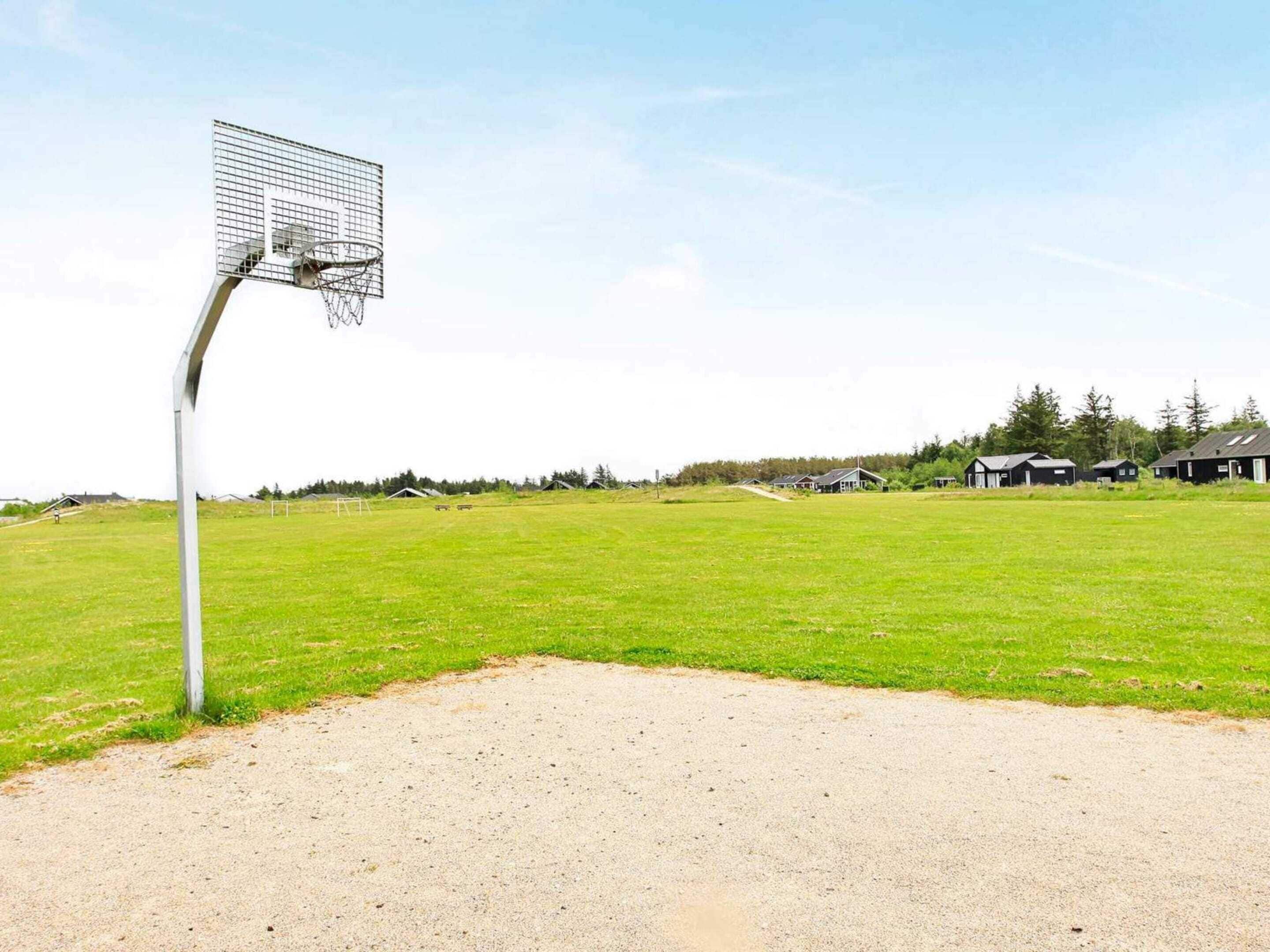 Una cancha de baloncesto con aro, rodeada de un campo de césped y casas al fondo.