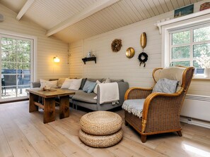 A cozy room with a wooden ceiling, a grey sofa, a wicker chair, a wooden table, and wicker ottomans.