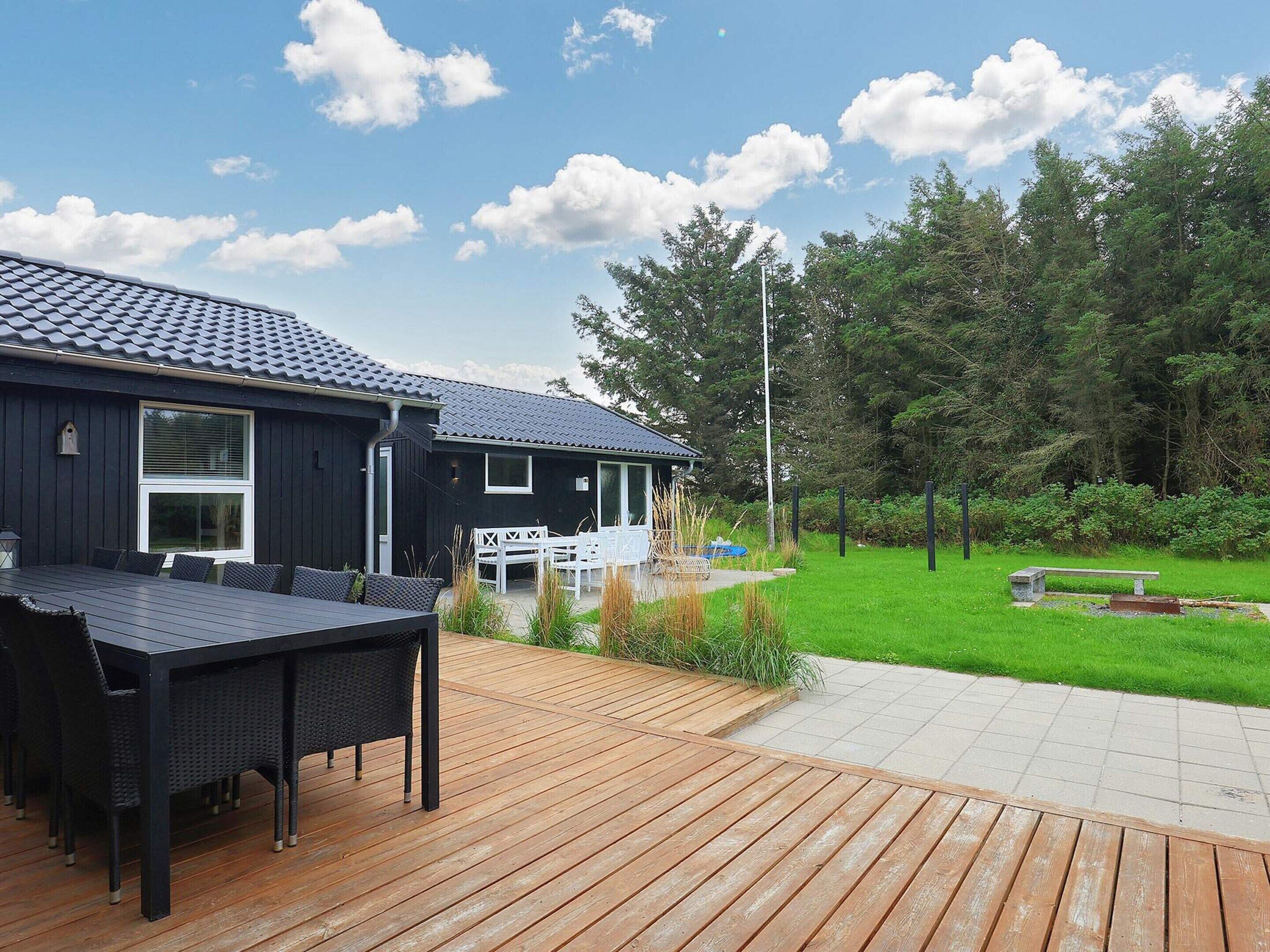 En træterrasse med spisebord og stole, en overdækket patio og udsigt til et hus med mørk facade og tegltag.